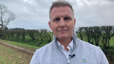 Chris Bailey, from The Air Ambulance Service, wearing a grey top and standing on land at Catthorpe, in Leicestershire, with a hedge line and trees in the background.