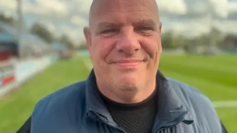 Karl Lindsay, in his 50s, who is bald and clean shaven, stands on a football pitch wearing a blue gilet over a black sweatshirt and smiles at the camera.