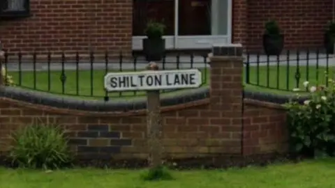 A road sign on the grass which reads Shilton Lane