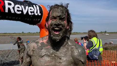 A man smiles at the camera covered completely in mud. His long black hair is similarly covered and he is not wearing a top. Other people covered in mud can be seen behind him as a photographer takes pictures.