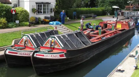 David Hawgood/Geograph A narrowboat painted in red with NUNEATON in white lettering on the front is lying alongside a much shorter craft similarly painted with the word BRIGHTON on the side. The boats have covered canopies at the front. There are people standing on the bank which is mostly grass with some stone steps up to a white waterside cottage.