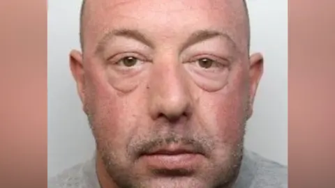 A close crop of a mugshot. A man with a pink face and short brown and grey stubble looks into the lens.