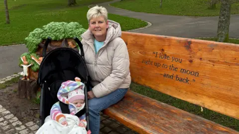 Roberta Carser, a woman with short, blonde hair, sits on a carved wooden bench with her hand on her granddaughter's buggy. She is wearing a padded beige jacket, blue jeans and a light blue t-shirt. Her granddaughter Torie is wearing a multi-coloured hooded jacket and a pink and white woolen hat. The bench features decorative carvings of trees and hares and it is engraved with a line from the book: "I love you right up to the moon and back."