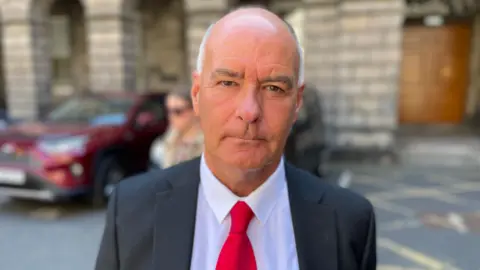 Tommy Sheridan standing outside the Court of Session - he is dressed in a black jacket, white shirt and red tie. He is mostly bald with white hair.