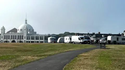 Caravans are parked across a mown lawn of grass, with a path running through it. Behind them is the Spanish City venue, an elaborate, white building with a dome roof.