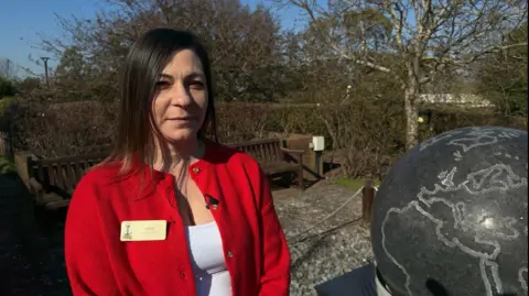 A woman in a red cardigan stands in a garden next to a model of a stone globe