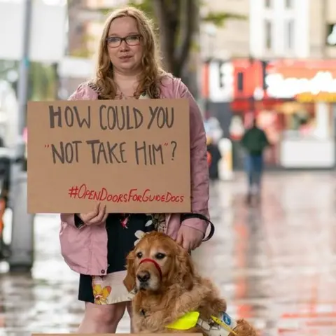 Guide Dogs Angharad Paget-Jones holding a placard saying 'how could you refuse him?' next to her guide dog Tudor