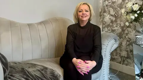 A photo of Mandy sitting on her sofa smiling. She has blonde hair and is wearing a black top and black trousers. 