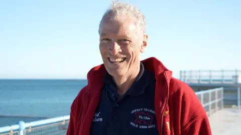 Chairperson of the International Marathon Swimming Hall of Fame Ned Denison, he is wearing a red hoody and a blue t shirt. he has short grey hair and is smilling.