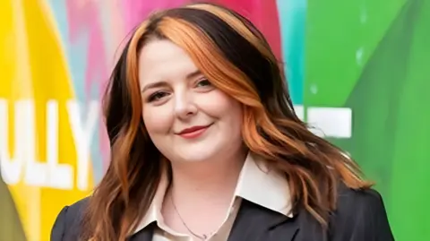 A woman with long brown hair with red highlights is wearing a dark jacket and standing in front of a slightly blurred multi-coloured hoarding.