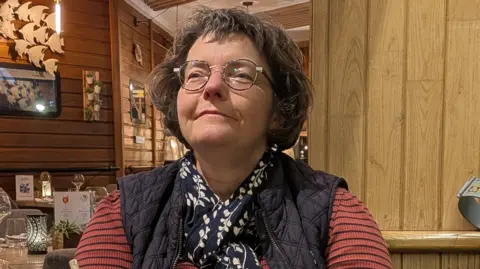 This picture shows a person seated at a table inside what looks like a cosy, warmly lit restaurant or café with a strong wooden interior. The space has wood-panelled walls, wooden ceiling slats and soft hanging lights that give the scene a relaxed, intimate feel. Decorative details are visible in the background, including framed items on the walls and small ornaments hanging from the ceiling.