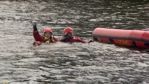 A red rubber inflatable boat is capsized. Two men in red dry suits, helmets and life jackets float in the water. One is waving as if calling for help.