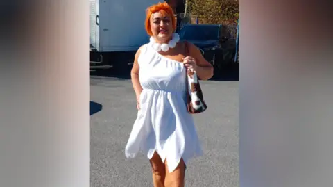 Lisa Hughes Woman wearing a white frayed dress and orange wig carrying a cow-hide-look bag