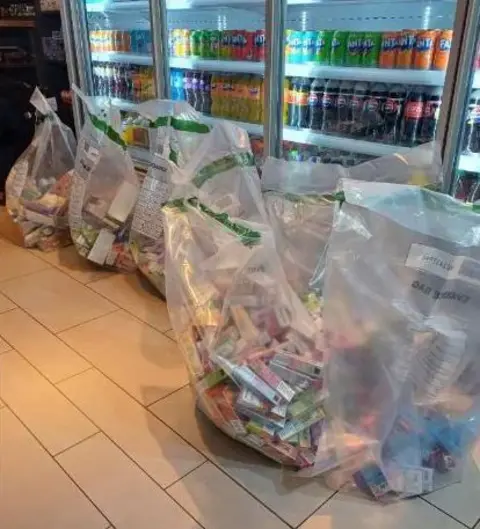 Nottinghamshire Police Six large see-through bags full of disposable vapes are sat on the floor of a shop which has drinks fridges behind them. 