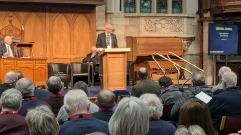 Presbyterian Church in Ireland/Chris Nelson Peter Gamble speaking at a podium on a stage. He has grey hair, wearing glasses, a dark blazer, white shirt and blue tie. Behind him is wood panelling. A man is sitting behind him, another man is sitting in an ornate style chair in the centre of the stage in front of a table. Blue carpet is on the stage. The backs of heads are in the audience. To the far right there is a screen reading: "General Council".