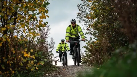 Merseyside Police Two police officers in hi-vis jackets and wearing cycling helmets ride e-bikes along a path. Shrubs and trees can be seen to either side of them. 