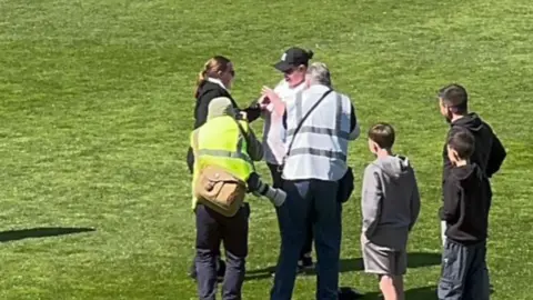 Contributed A woman places a ring on another woman's finger in front of two photographers on a football pitch. One woman wears a black jumper with sunglasses and black trousers. She has dark hair that is tied back. The other woman wears a black cap, sunglasses, a pink T-shirt and black trousers.