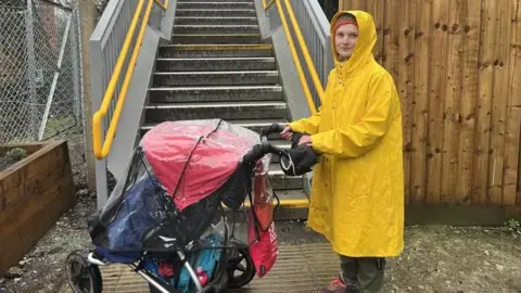 A woman in a yellow coat holding a pushchair. She is stood at the bottom of some metal stairs.