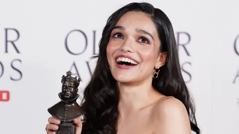 Picture of Rachel Zegler smiling and holding her Olivier award in front of a white board with the text 'Olivier awards' on it.