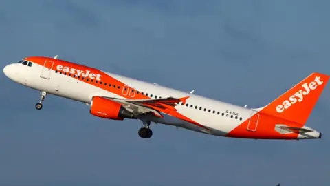 An EasyJet Airbus A320 passenger aircraft climbs after take-off against a clear blue sky. The aircraft is painted in company's distinctive orange-and-white livery.
