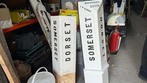 Two white signs on the floor, standing up, saying "Dorset" and "Somerset". They are standing up in a garage.