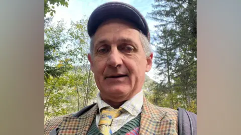 Robert Elwes A selfie-style picture of Robert Elwes, who is wearing a flat cap, checked jacket and a multi-coloured jumper with yellow and blue tie. He is standing outside surrounded by trees.