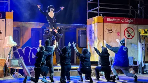 Newcastle United mascot, Monty Magpie, is held aloft by two men on stage. Six other actors are on their knees holding their arms up towards him in praise.