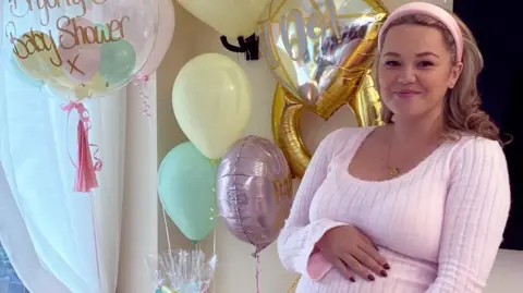 Russo family Bryony Russo wearing pink lounge clothes and a headband. She is smiling and holding her baby bump at a baby shower, with lots of balloons behind her.