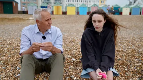 A male reporter with grey hair wearing a blue shirt sitting on a shingle beach with a young woman with long dark hair who is wearing a black sweatshirt with a row of colourful beach huts in the background.