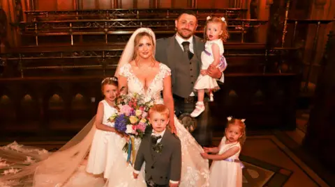 McGeoch Family A wedding portrait of a couple and their four children. The bride is wearing a white wedding dress, while the groom is in a grey suit and a kilt. Their four children are gathered around them. 