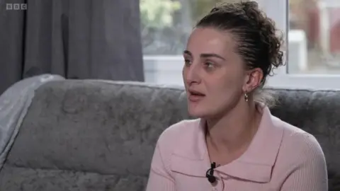 Cooper sitting on a sofa in a living room. She is turned slightly to the side, speaking or listening to someone off‑camera. She is wearing a pale pink top with a collar and has a small microphone clipped to the front. Her hair is pulled back, and she is wearing small hoop earrings. Behind her, grey curtains and a window with soft daylight coming through can be seen, along with a grey textured sofa.