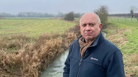 A man in a blue fleece is standing in front of a small stream and surrounded by fields.