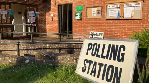 A black and white sign reads 'polling station' outside the entrance to Shoreham Village Hall. There is bunting above the door and signs directing votes into the building. There is no one in the photo.