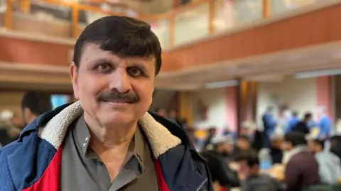 Sarfaraz is wearing a winter coat stands in the foreground of a large indoor hall. Behind them, many people are seated at tables, engaged in conversation or eating, with the upper level of the hall visible in the background.