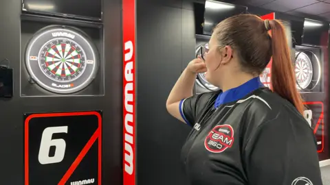 Lola throws a dart at a dartboard at the new Hangar61 venue. She has long hair tied back in a ponytail and wears a black team shirt.