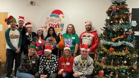 Stripey Stork Eleven people in two rows wearing Christmas jumpers and red Santa hats. They are next to a decorated Christmas tree and a sign that reads Santa Stork.