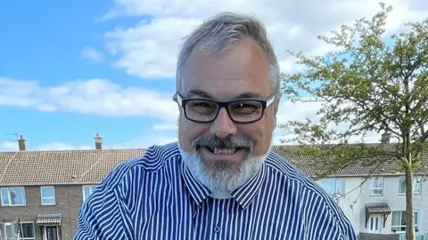 Stephen Doherty A man is pictured outside smiling. He has white hair and beard. He is wearing black glasses and a blue shirt with white strips. Terraced houses can be seen in the background as can a tree.