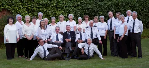 ESSEX POLICE COMMUNITY CHOIR 26 members of the choir, most wearing white tops.  Most are standing on a lawn with some sitting down.  Green foliage in the background.