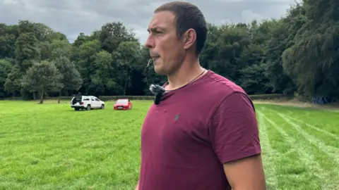 A man stood in a field with trees behind him, he is blowing into a whistle and wearing a pink top. 