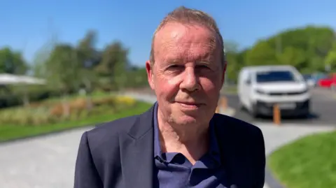 A man in his seventies wearing a dark shirt and jacket with a ruddy complexion.