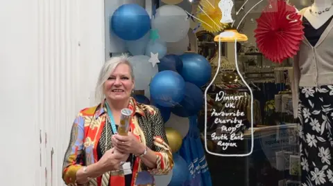 Adrian Harms/BBC Branch manager Julie Bunting holds a streamer in front of her standing in front of the shop window with decorations and clothes seen in the background