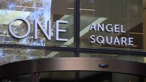 White letters saying "An Angel's Square" in glass windows above a brown-framed revolving door to a public building.