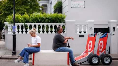 EPA/Shutterstock A man and woman sitting on either side of a large white anti-terrorism concrete block on Portobello Road. In the background is a white wall with a balustrade with greenery.