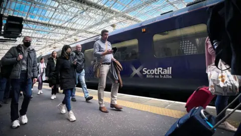 Commuters walk along a train platform next to a dark blue train which reads 'ScotRail' in white writing next to a saltire. A glass roof above the station shows a blue sky.