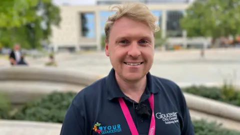A man with light blond hair smiles as he stands in a city centre square. He is wearing a navy Gloucester City Council polo shirt and a pink lanyard. People can be seen chatting in the square behind, where there are trees and a big, modern university building.