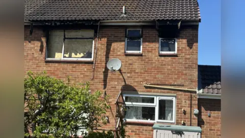 Melissa Head A classic 60s style semi-detached house built with red bricks. The top is blackened from the fire, and the windows on the first floor are boarded up and severely damaged.