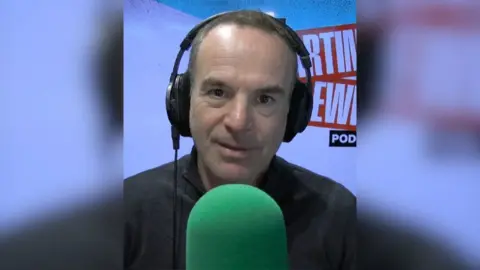 Martin Lewis wears a black top and headphones and sits in front of a green microphone