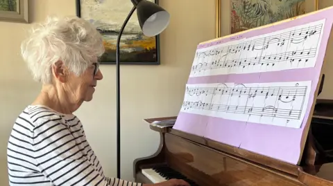 A woman sits in front of a piano reading large sheet music as she plays. She has short grey hair and wears a white long sleeved top with black stripes on it. She is also wearing glasses. 