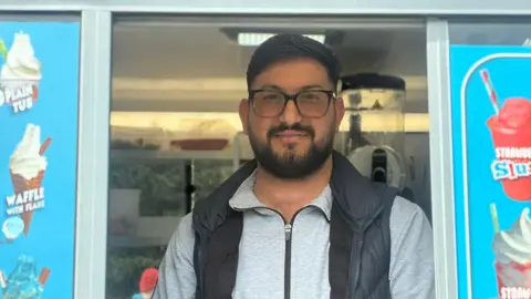 Matt Precey/BBC Man with glasses and a beard wearing a gilet. He is standing in the serving hatch of an ice cream van.