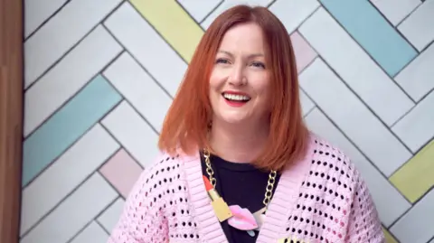 Kirsty Sedgman Dr Sedgman stands in front of pastel-coloured tiles. She has shoulder length copper hair and smiles at the camera. She wears a pink cardigan, with a black top underneath and a chunky necklace. 